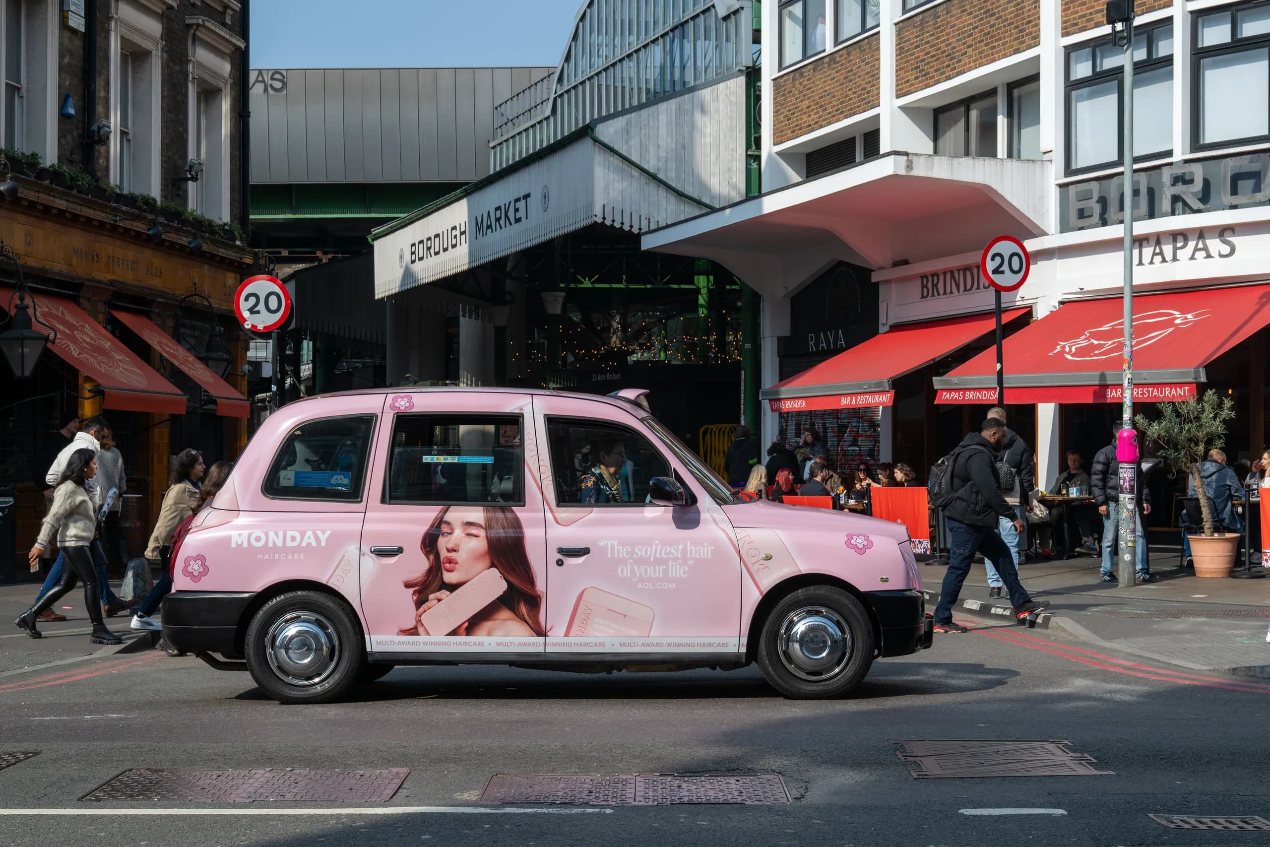 Taxi in pink Monday Haircare livery on a busy street, red shop awnings behind