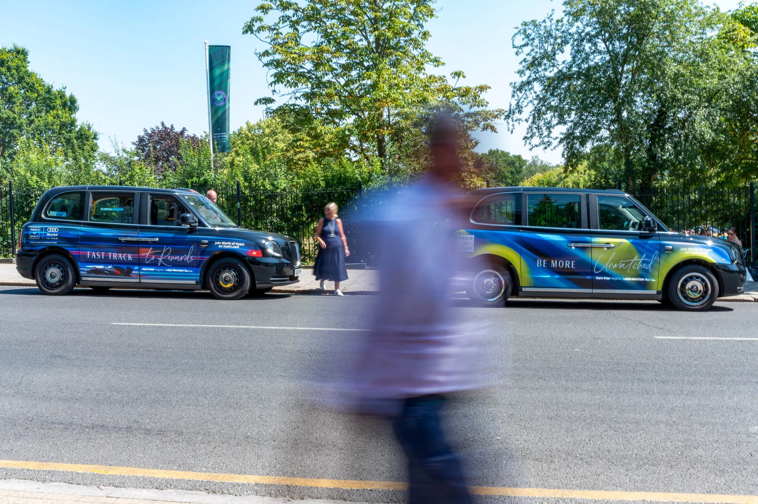 Two taxis with World of Hyatt advertising on a tree-lined road in Wimbledon, a passer-by blurred in front